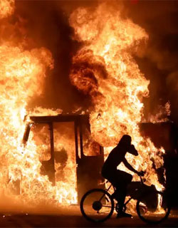 Man on bicycle riding past building in flames, Kenosha WI